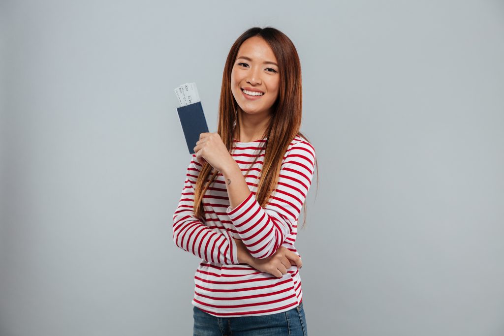 Smiling asian woman in sweater holding passport with tickets and looking camera over gray background